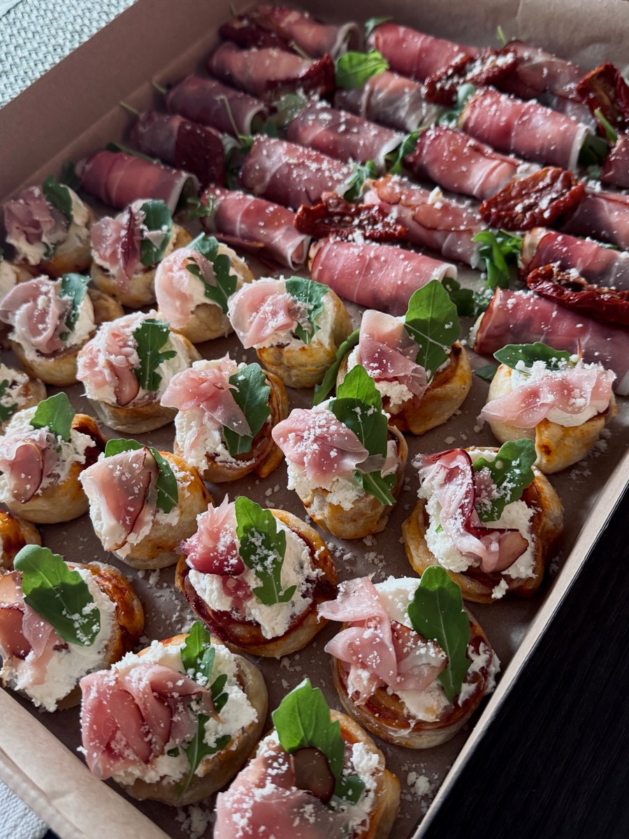 Tartinki z ricottą, rukolą i szynką dojrzewającą, w tle roladki z prosciutto z suszonym pomidorem