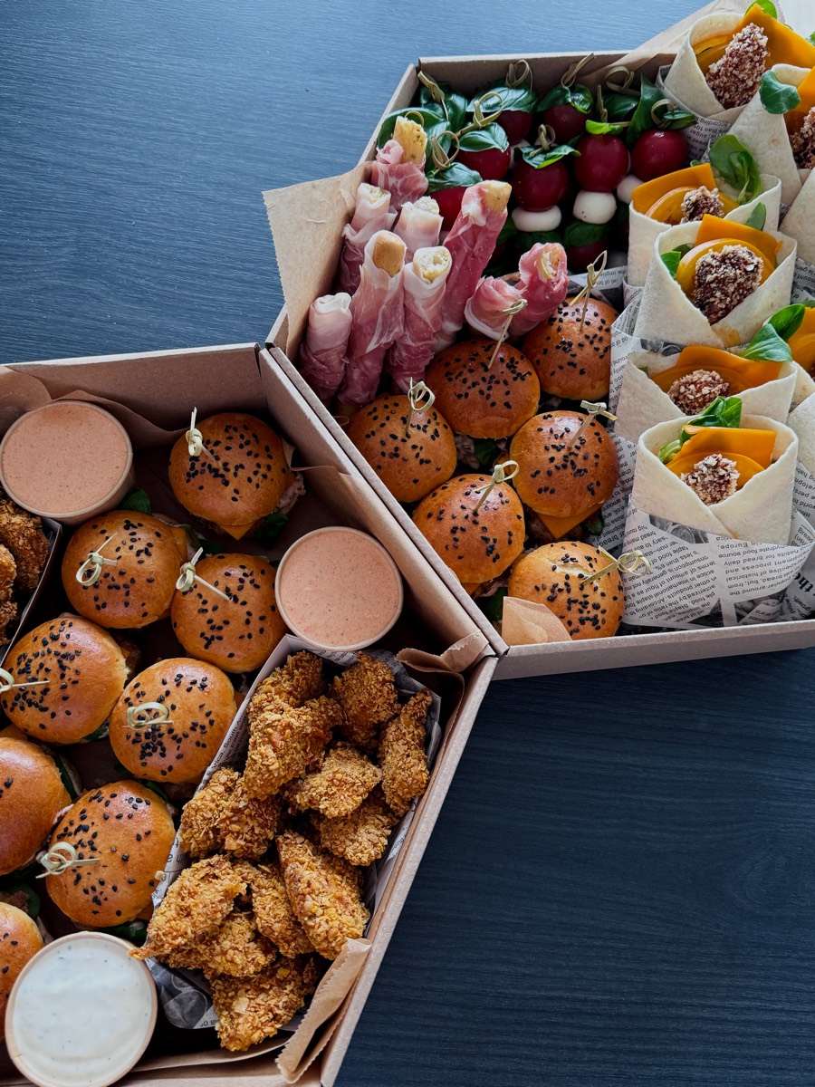 Trzy boxy obok siebie: mini burgery z czarnuszką, nuggetsy z kurczaka w panko z dwoma sosami, koreczki caprese, grissini owinięte szynką dojrzewającą i wrapy z panierowanym kurczakiem
